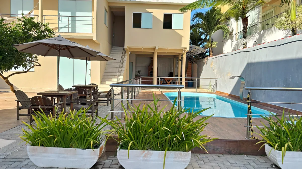 Pátio do Residencial Maria Rita: piscina, mesa com guarda-sol e a casa ao fundo, em proporção 16 por 9